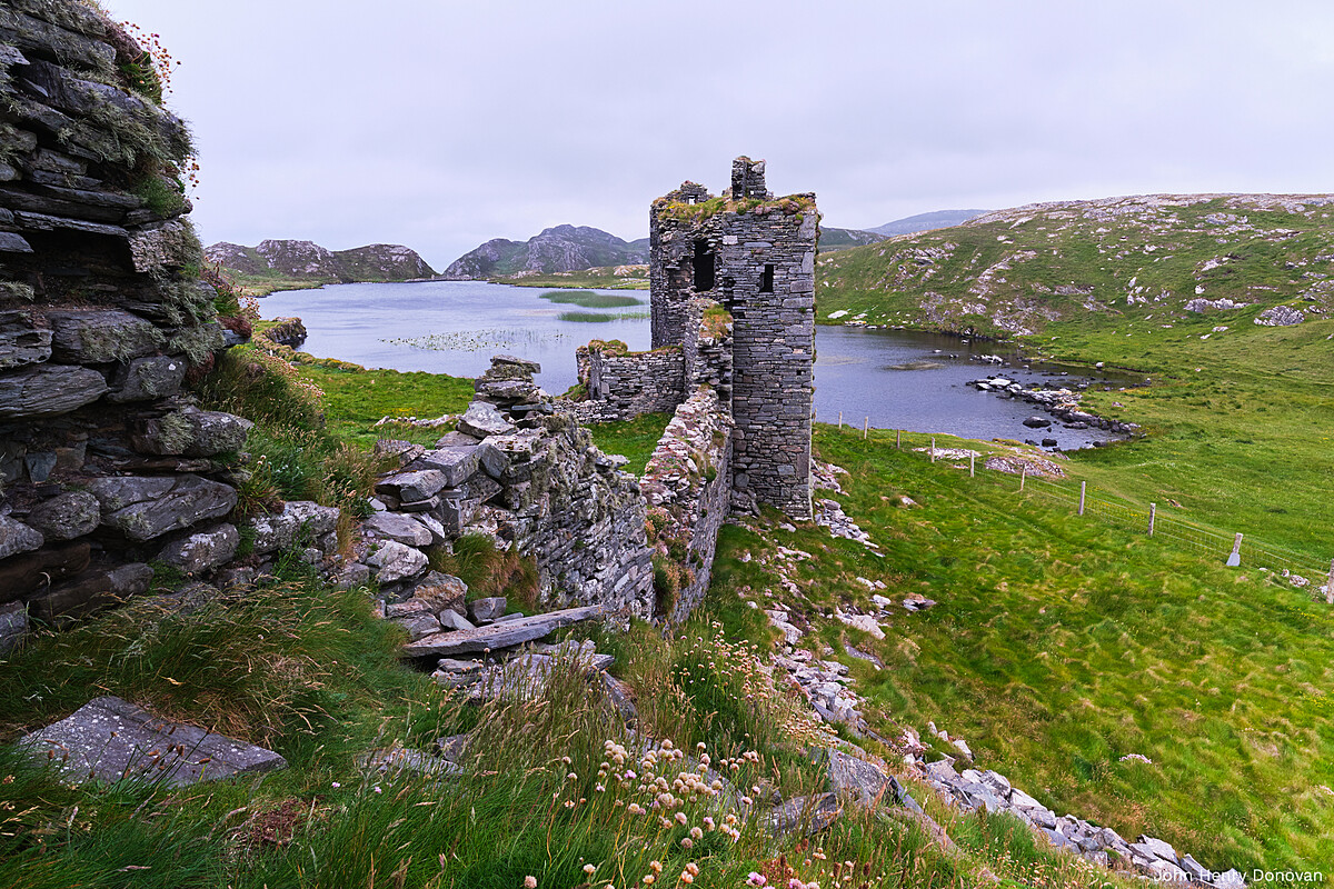A weathered stone ruin stands on a grassy hill overlooking a tranquil lake surrounded by rolling hills. The sky above is overcast, casting a muted light over the landscape. Wildflowers and rocks dot the foreground, enhancing the scene's natural beauty.