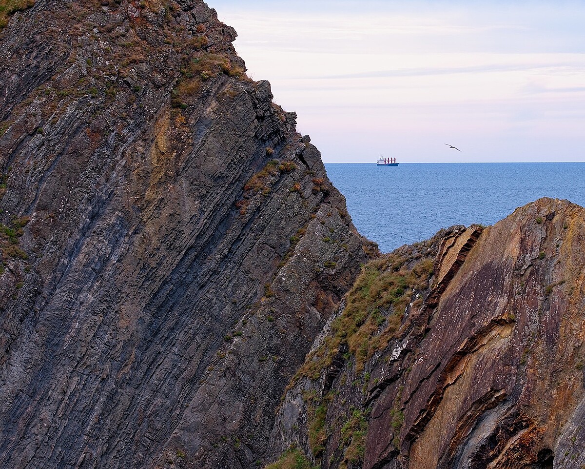 A large rock formation juts out into the ocean from both sides of the image, partially framing a cargo ship in the distance. Above the calm blue water, the sky transitions from a pale blue to soft pink hues, with a single bird flying near the rocks.