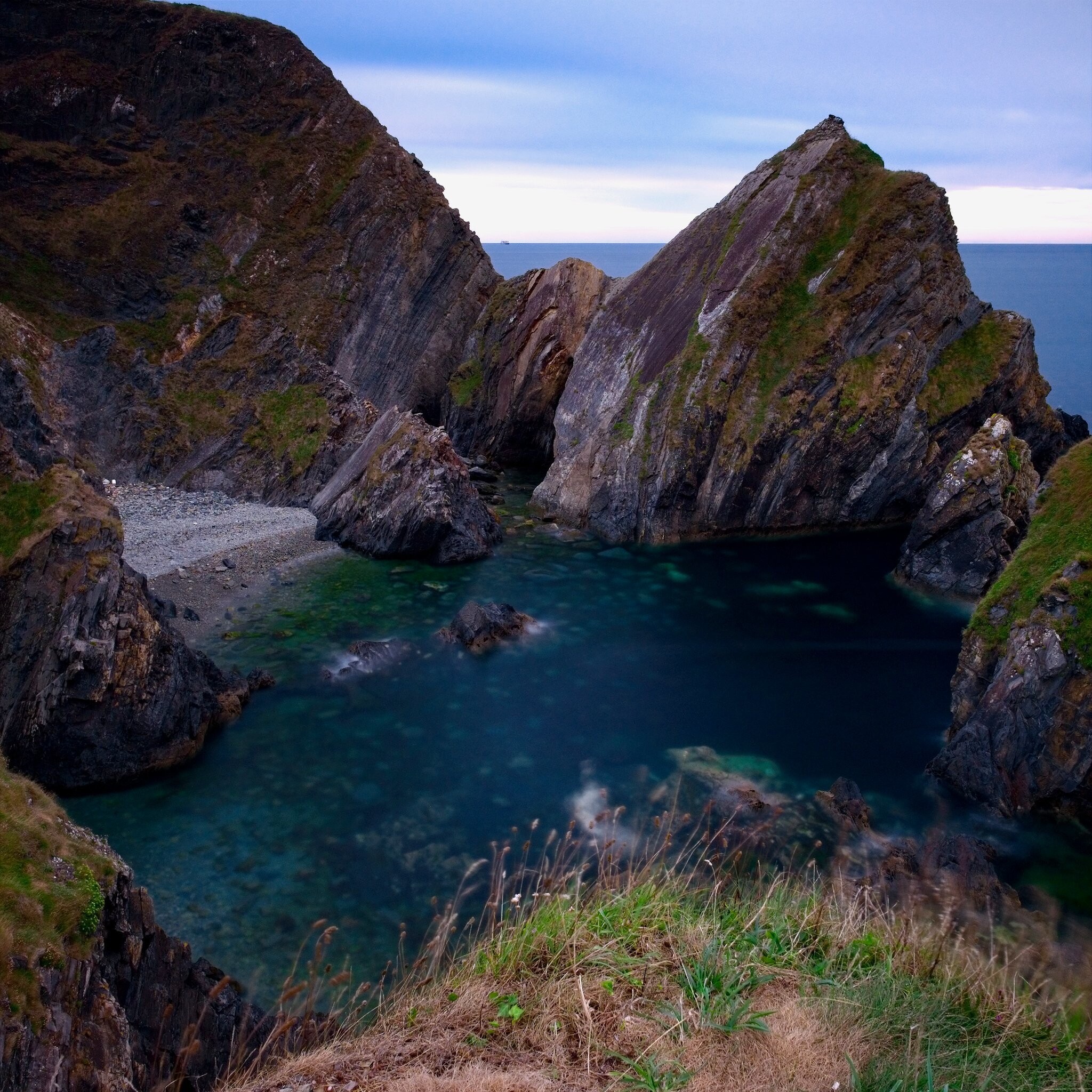 View of a coastal inlet with rugged cliffs on either side and clear blue-green water below. The steep cliffs have patches of mossy vegetation. There is a small, pebbly beach at the base of the cliffs. Sky is overcast, suggesting cool or overcast weather.