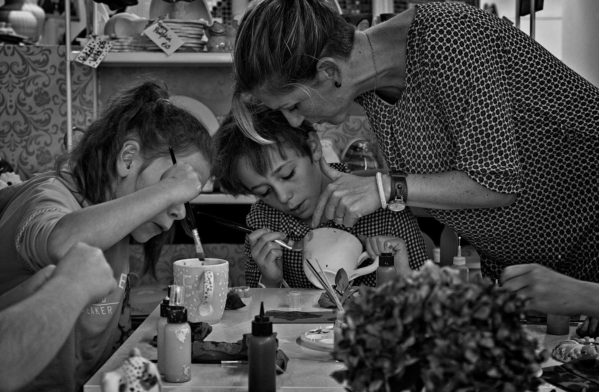 A black-and-white photo depicts a group of children engaged in painting ceramics at a table. An adult is closely assisting a child holding a paintbrush. Bottles of paint and ceramic items are scattered on the table, with decorative backgrounds visible.