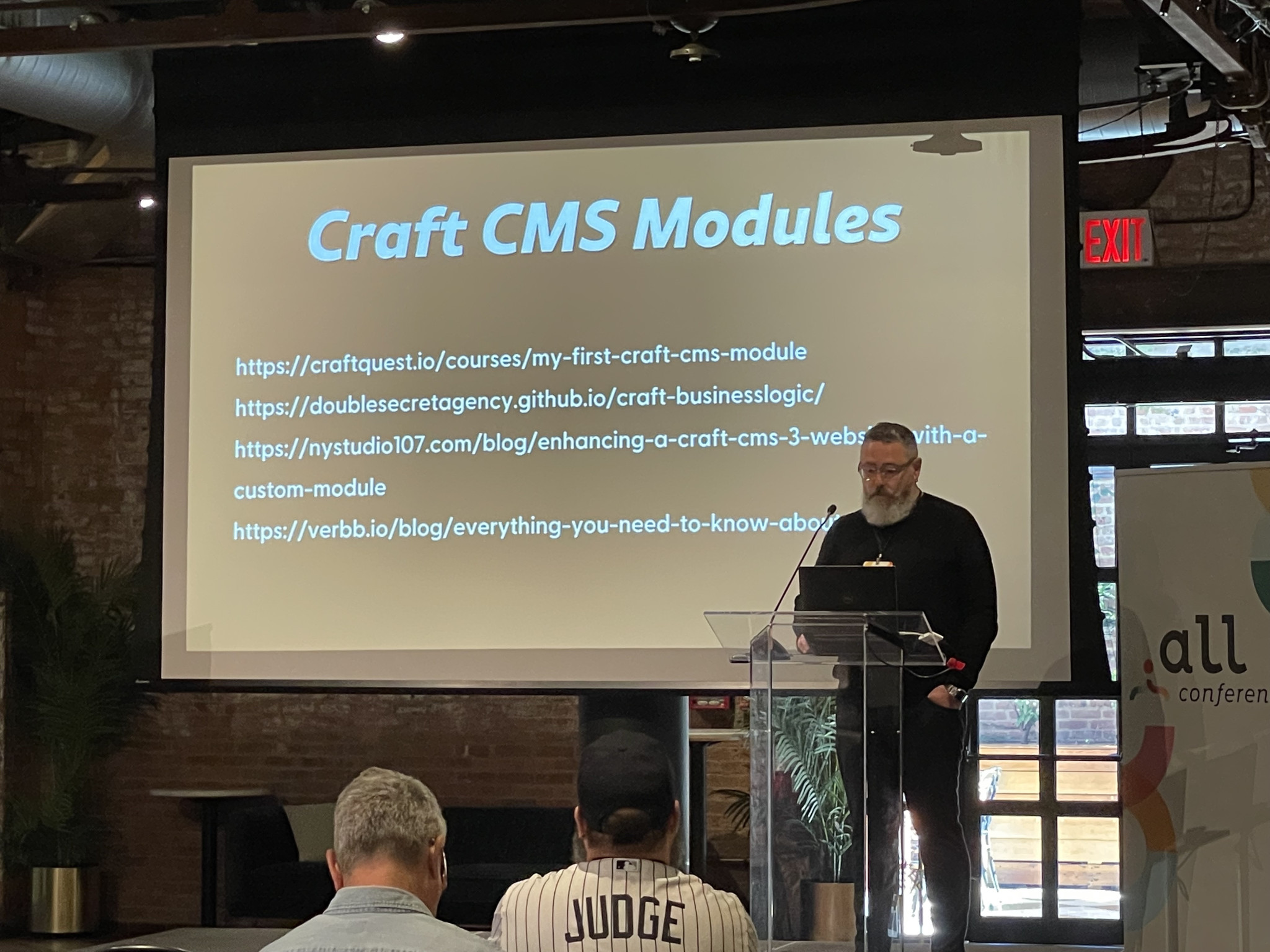 A man is giving a presentation on Craft CMS Modules at a conference. The slide behind him displays several URLs for resources related to Craft CMS Modules. Attendees are seated and attentively listening to the speaker.