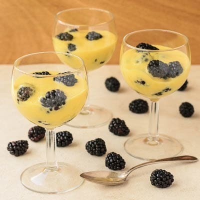 Three glasses of custard topped with blackberries on a light wood surface, accompanied by extra blackberries and a spoon.