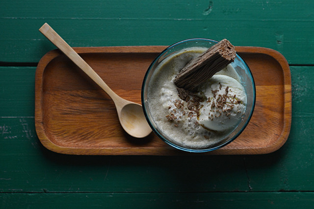 A glass bowl with vanilla affogato, topped with a flake chocolate bar and grated chocolate, sits on a wooden tray with a wooden spoon against a green wooden background.