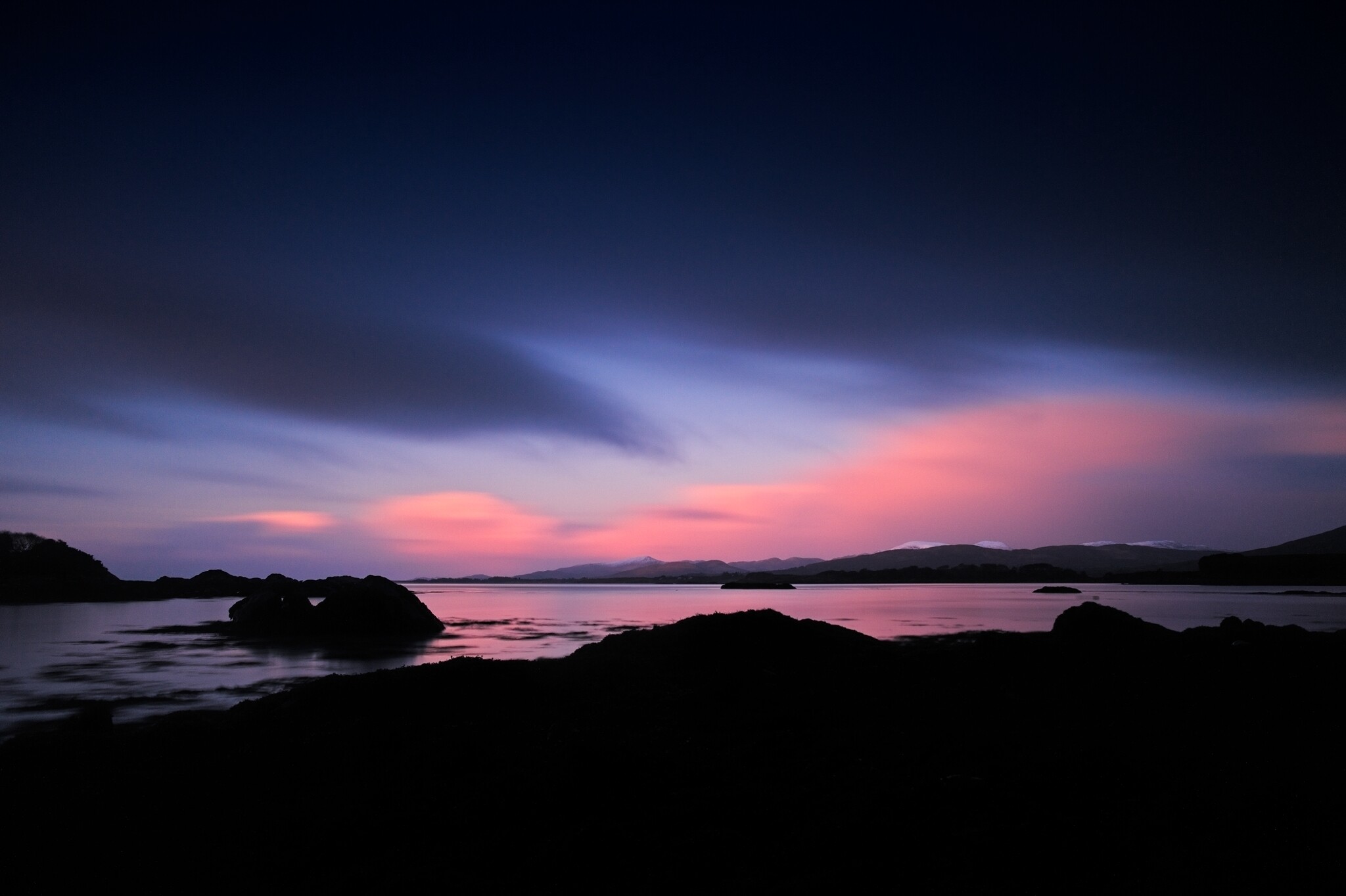 A tranquil coastal scene at dusk with a dark foreground contrasting a colorful sky. The sky transitions from deep blue to pink and purple hues, reflected in the calm waters. Rock formations and distant mountains are silhouetted against the horizon.