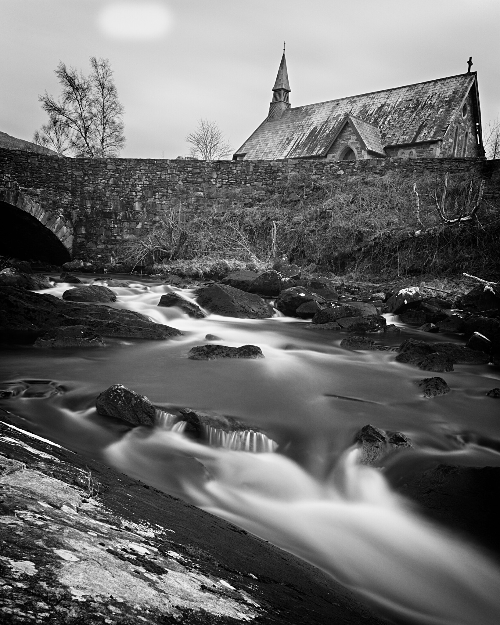 Black and white photograph of a serene landscape featuring a flowing stream with rocks in the foreground, a stone bridge above the stream, and a rustic church with a steeple in the background.