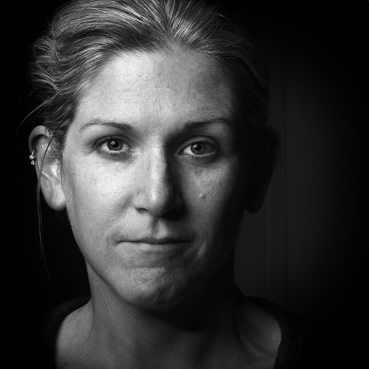 Black and white close-up portrait of a person with light-colored hair pulled back. They have a neutral expression, and their face is centrally framed against a dark background. Light and shadow accentuate the facial features. An earring is visible in the left ear.