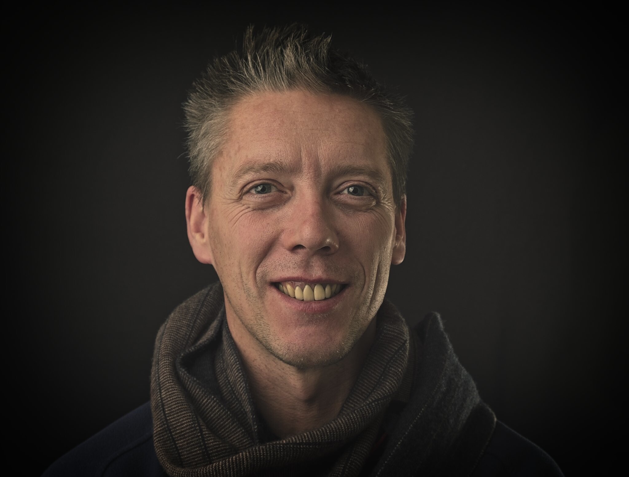 A man with short gray and black hair is smiling at the camera. He is wearing a dark-colored jacket and a brown scarf. The background is dark.