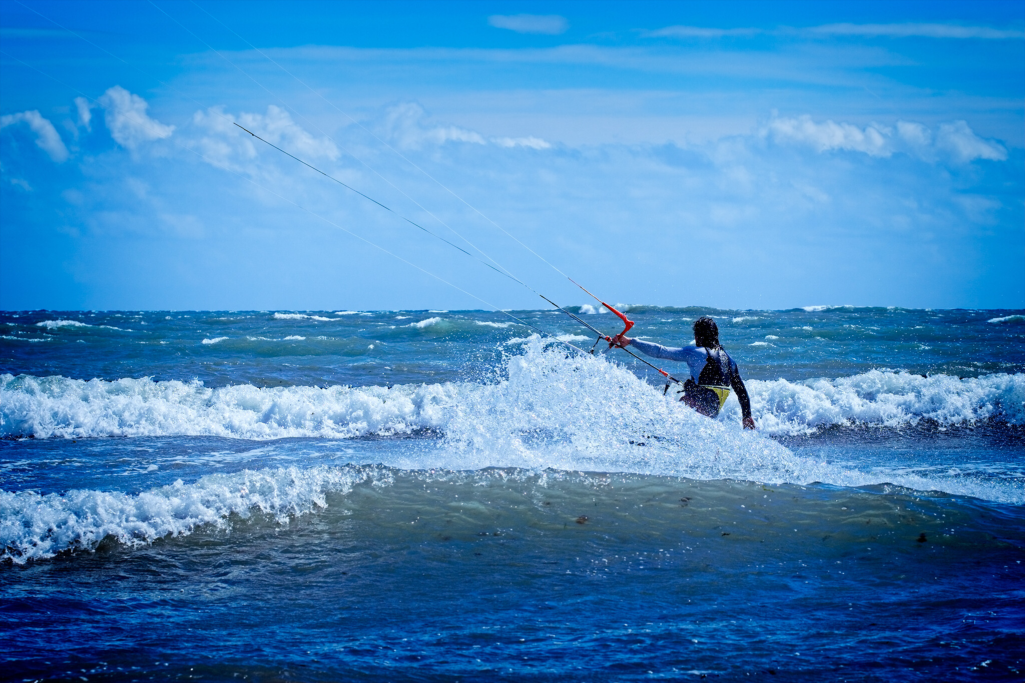 A person engages in kitesurfing on a body of water. The individual is holding onto the kite's control bar as waves crash around them. The sky above is mostly clear with scattered clouds.