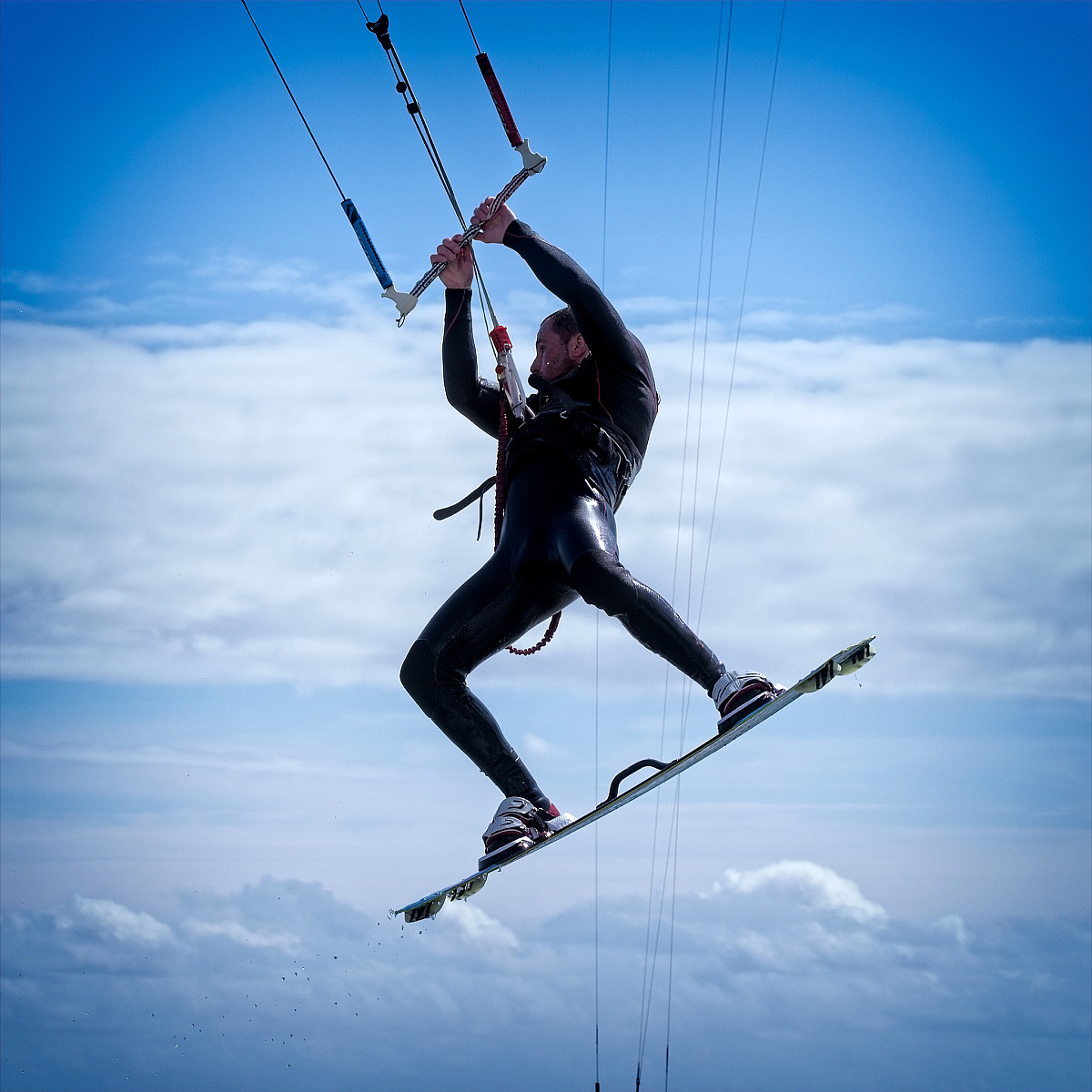 A person wearing a black wetsuit is kiteboarding against a backdrop of a cloudy sky. They are airborne, holding onto the kite control bar with both hands, and their legs are bent slightly at the knees. The person appears to be performing a jump or trick.