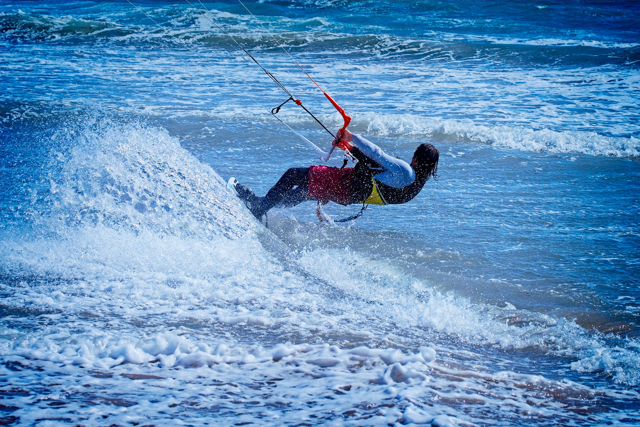 A person kiteboarding in the ocean near the shore, holding onto the kite control bar while skimming over the water with waves in the background. The individual is dressed in a wetsuit and other protective gear. The sea water is splashing around them.