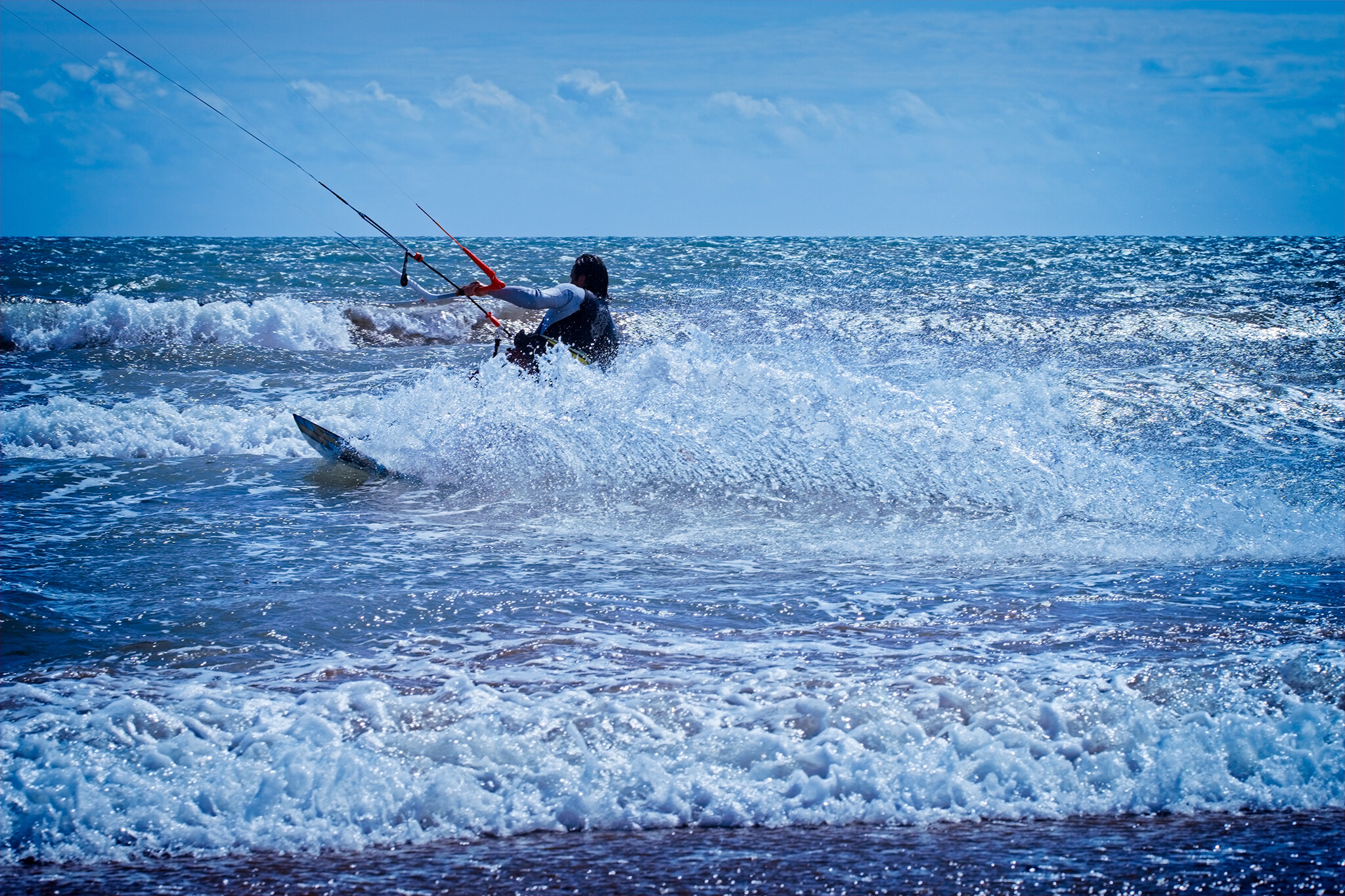 A person wearing a life vest is kite surfing in the ocean. They are maneuvering their board through the waves, with the kite lines visible in the background. The ocean is choppy with small waves, and the sky is bright and clear.