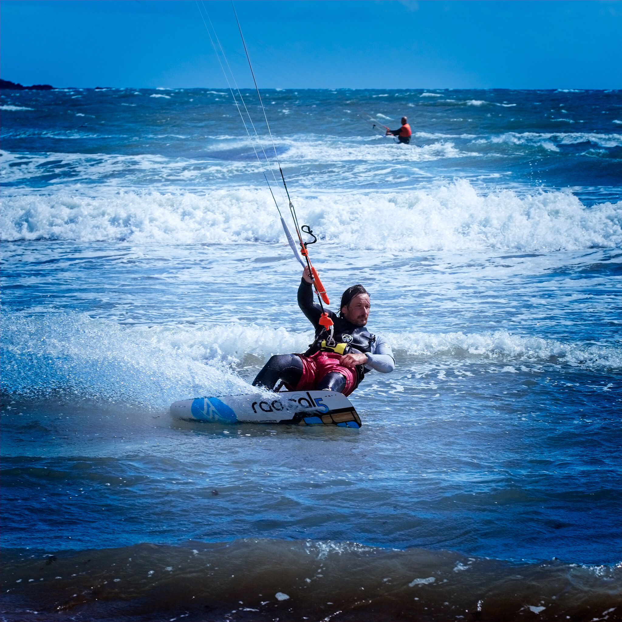 A person is kiteboarding on a wavy sea, holding onto the kite's control bar. The individual is wearing a wetsuit and a life vest. Another person can be seen kiteboarding in the background. The sky is clear with a few clouds.