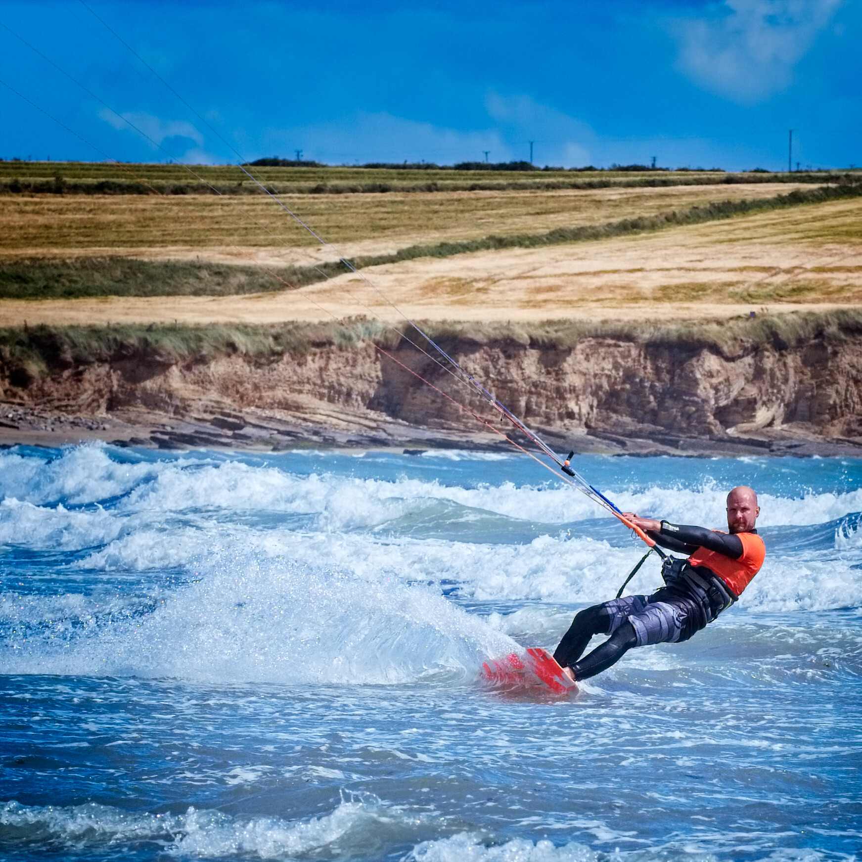 A person in a wetsuit is kiteboarding on the ocean with waves crashing around them. The person is grasping a control bar connected to a kite while gliding over the water on a kiteboard. The shoreline with grassy fields and power lines is visible in the background.