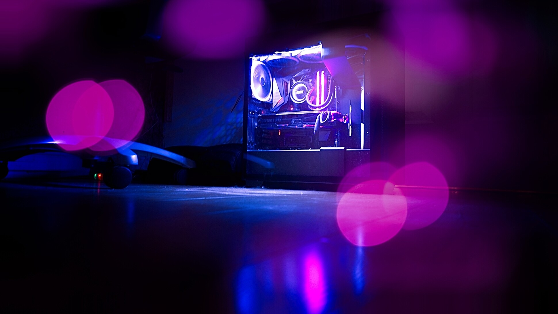 A desktop computer with a transparent side panel displaying internal components illuminated by purple lighting. The foreground and background are out of focus, with bokeh effect purple lights.