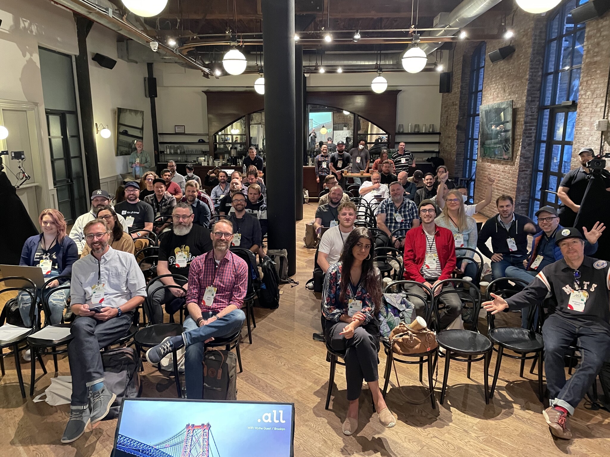 A group of people are seated in rows facing the front of a room, appearing to be at an event or presentation. Some individuals are smiling and waving at the camera. The setting includes industrial-style lighting, exposed brick walls, and a wooden ceiling.