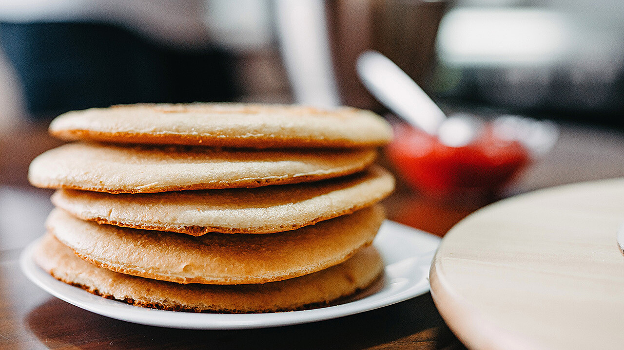 A stack of golden-brown pancakes is placed on a white plate. In the background, there is a blurred image of a person standing and a small bowl containing a red condiment, likely jam or syrup, with a spoon sticking out.