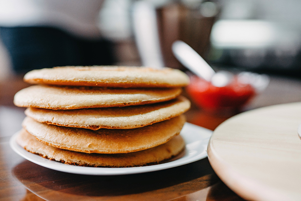 A stack of golden-brown pancakes is placed on a white plate. In the background, there is a blurred image of a person standing and a small bowl containing a red condiment, likely jam or syrup, with a spoon sticking out.