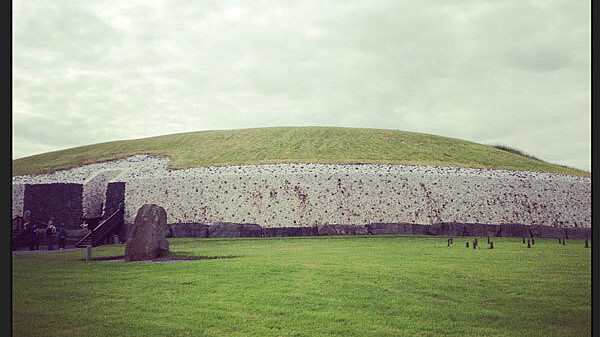 A large, grassy mound stands under a cloudy sky. The mound is partially faced with white stone with a dark entrance area visible to the left. A large standing stone is positioned in front of the mound on a well-kept lawn.