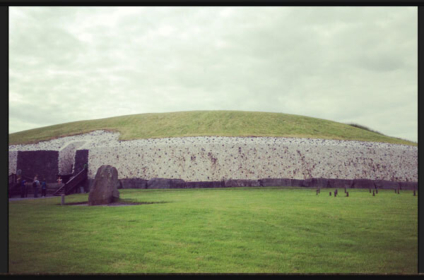 A large, grassy mound stands under a cloudy sky. The mound is partially faced with white stone with a dark entrance area visible to the left. A large standing stone is positioned in front of the mound on a well-kept lawn.