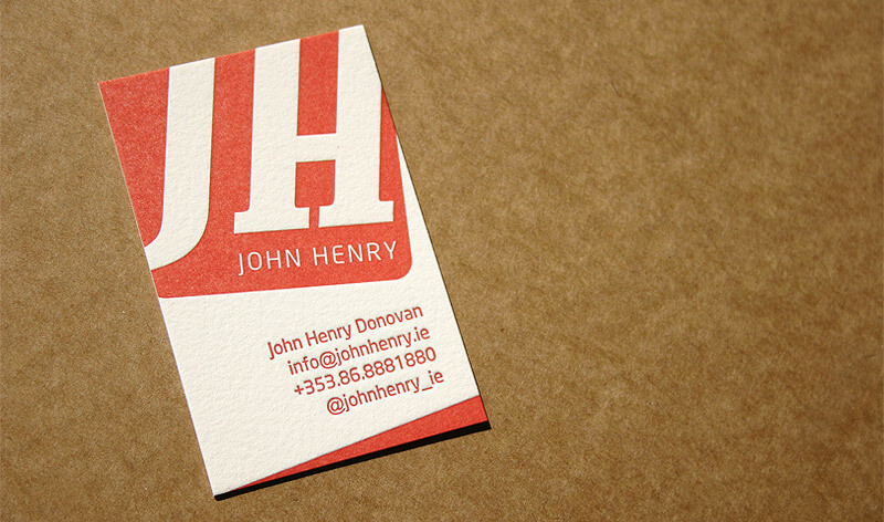 A business card lays on a brown surface. It features the large initials "JH" in red at the top, and below it the name "John Henry" in white. The card includes contact details: Email, phone number, full name (John Henry Donovan), and a social media handle.