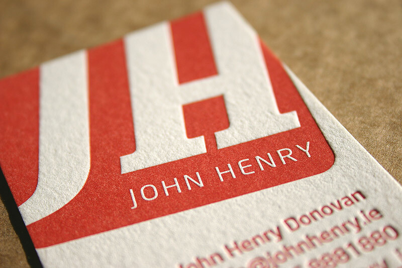 Close-up of a business card featuring large, bold red letters “JH” with the name “John Henry” written beneath in smaller white text. The card has textured paper and is angled slightly on a brown surface. Additional contact details are partially visible.