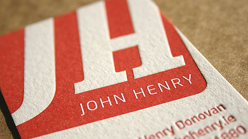 Close-up of a business card featuring large, bold red letters “JH” with the name “John Henry” written beneath in smaller white text. The card has textured paper and is angled slightly on a brown surface. Additional contact details are partially visible.