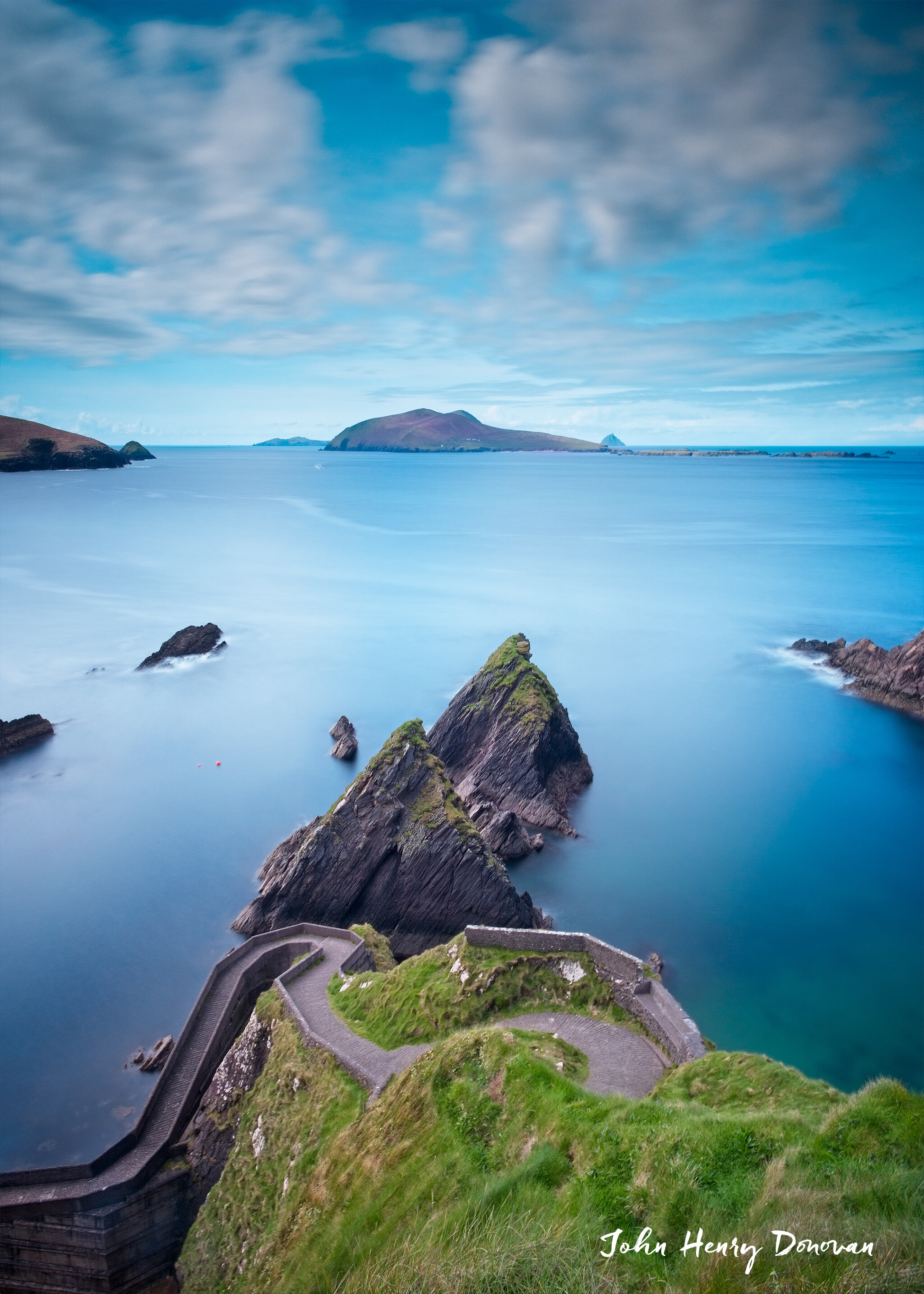 A scenic view of a curved path leading down to the ocean, with grassy cliffs and jagged rock formations. The calm sea extends to the horizon under a partly cloudy sky. An island is visible in the distance. "John Henry Donovan" is written at the bottom right corner.
