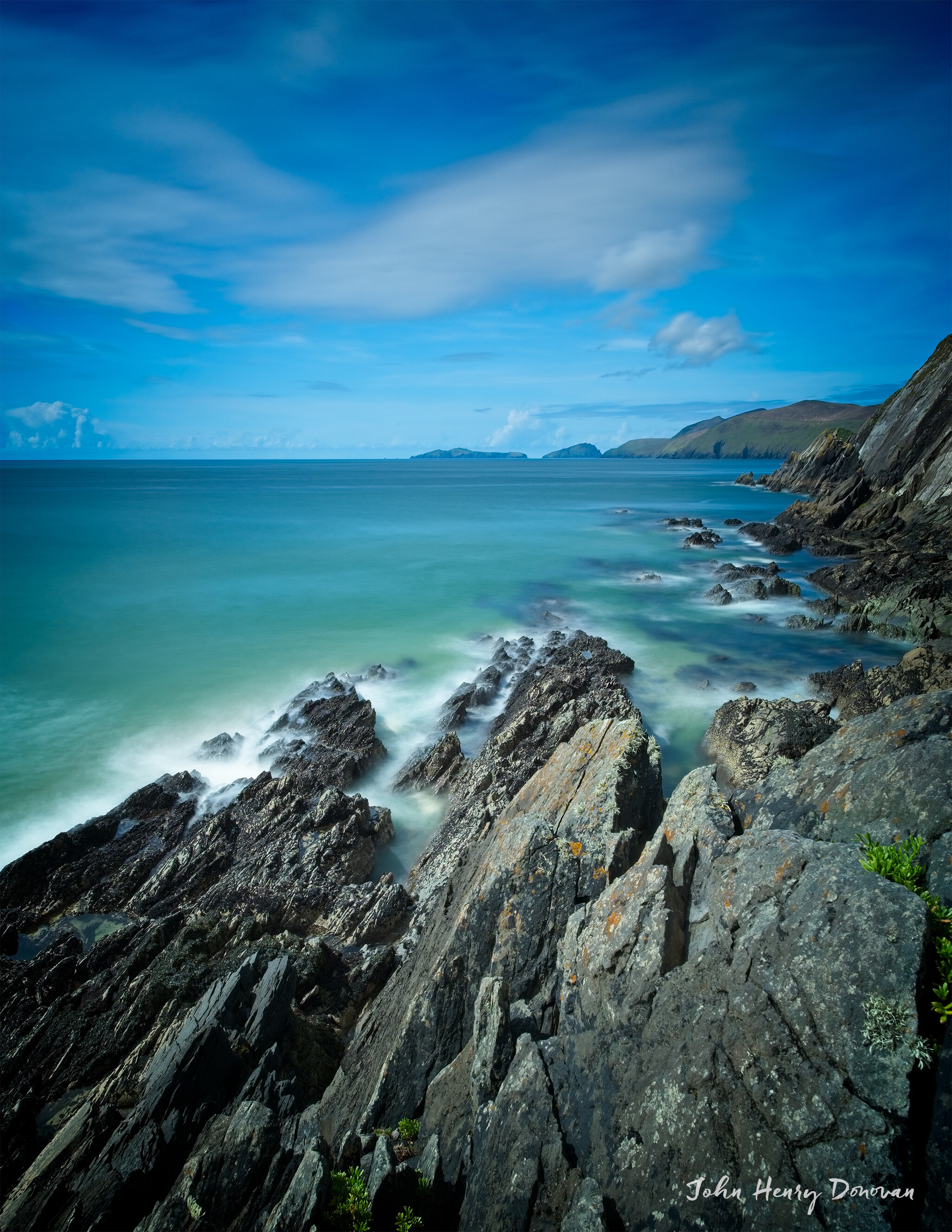 A rocky coastline under a blue sky with soft, wispy clouds. The ocean waves crash gently against the rocks, creating a misty effect. In the distance, green hills slope towards the sea. The image is signed "John Henry Donovan" in the bottom right corner.
