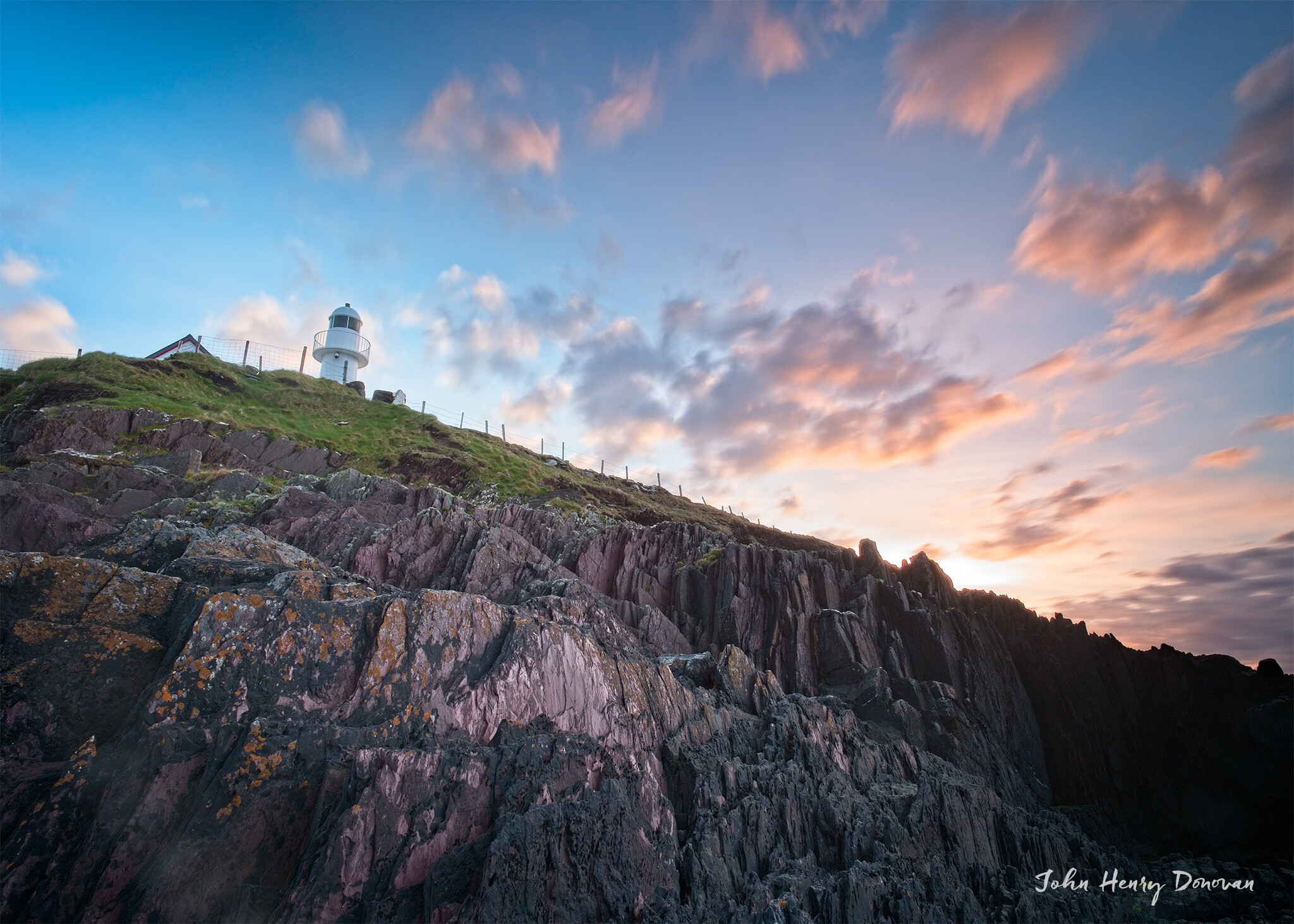 A white lighthouse stands atop a rocky hill under a sky with scattered clouds at sunrise or sunset. The rocks appear rugged and steep. Green grass is visible near the base of the lighthouse. The signature "John Henry Donovan" is at the bottom right.
