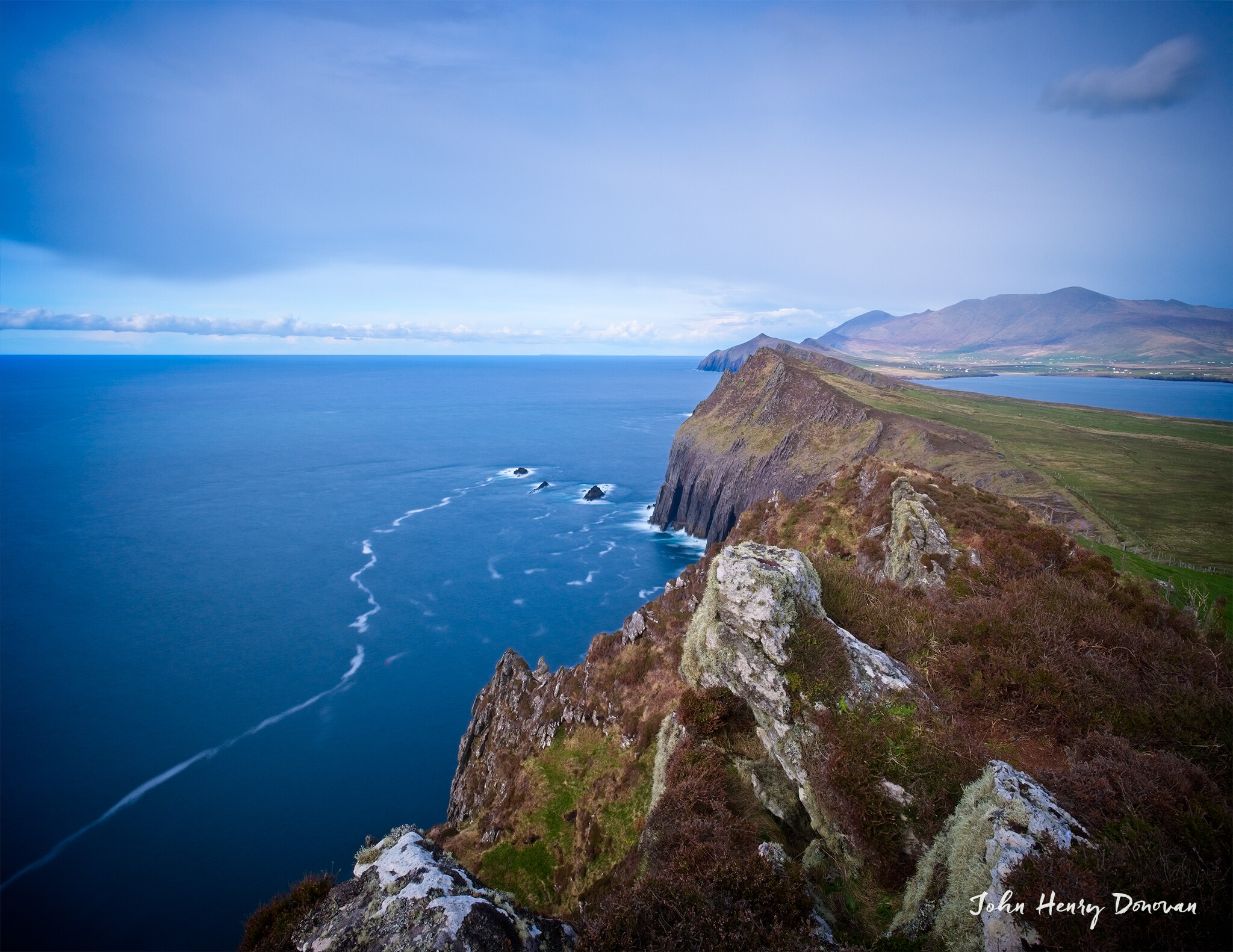A coastal cliffside view with rugged terrain overlooking a vast blue ocean under a clear sky. Below, waves form white streaks. In the distance, the cliffs extend towards rolling hills and a hazy horizon.