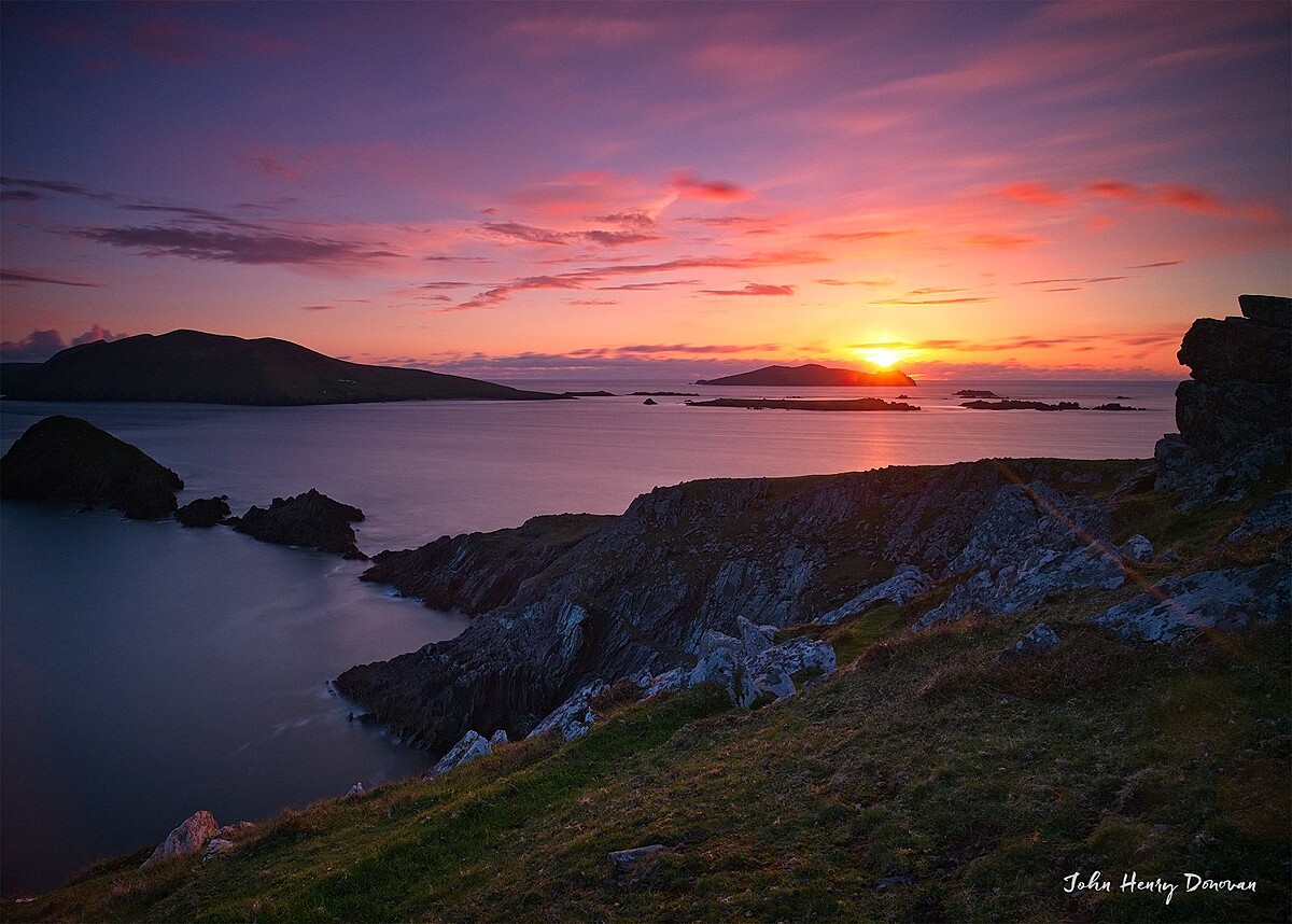 A vibrant sunset over a rugged coastal landscape. The sky is a mix of orange, pink, and purple hues. Dark, jagged rocks and calm waters are in the foreground, with distant islands silhouetted against the horizon.