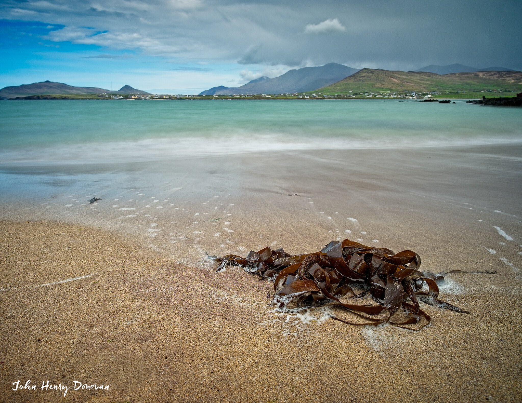 A beach with wet sand and seaweed in the foreground, waves gently washing up. In the middle ground, clear turquoise water extends to the horizon. In the background, mountains and a cloudy sky. A photo credit to "John Henry Donovan" is visible in the lower left corner.