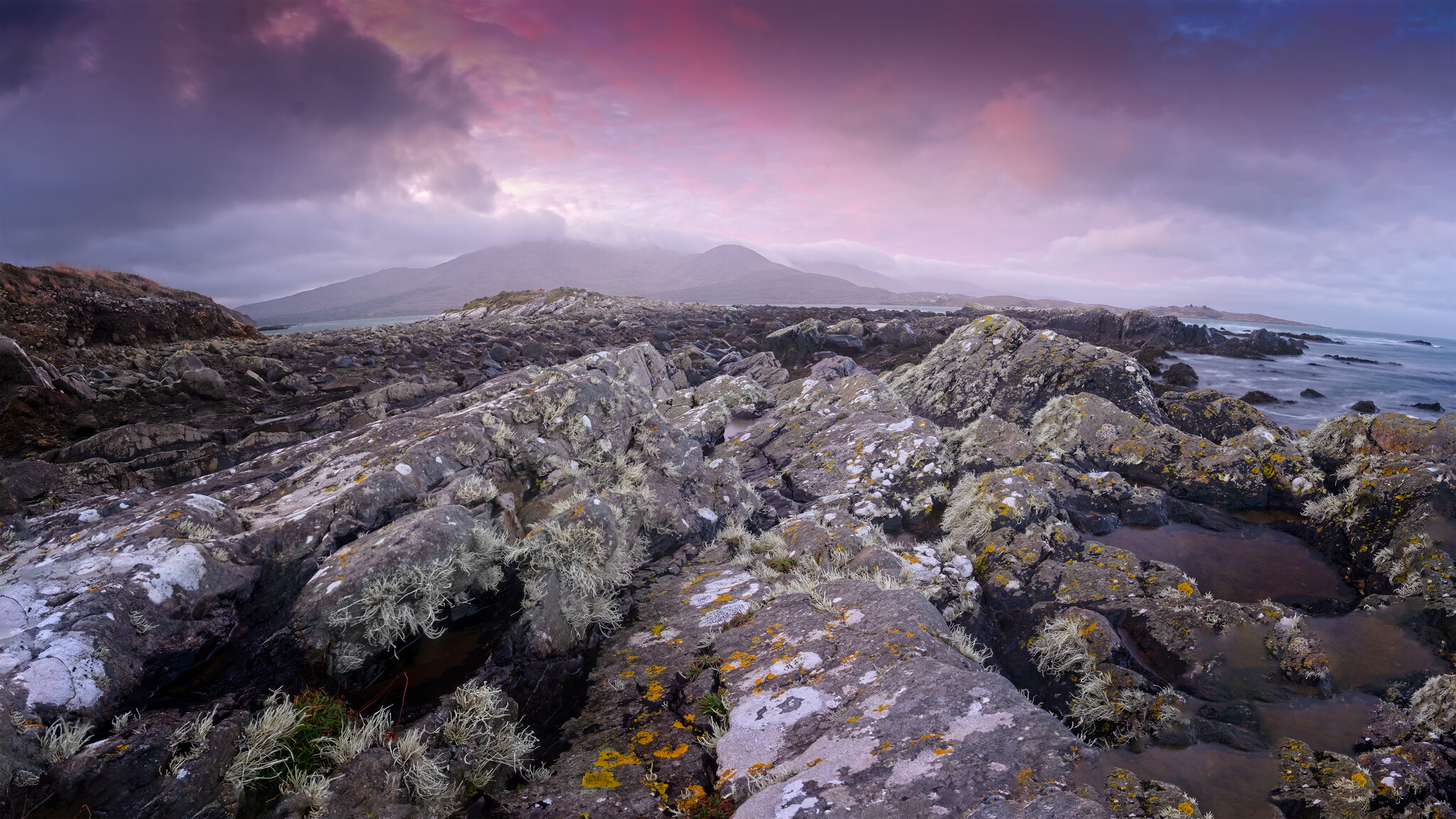 A rocky coastal landscape with rugged terrain covered in patches of lichen and moss. The ocean borders the rocks on the right, while a mountainous backdrop is visible under a purple and pink cloudy sky. The scene is tranquil and natural.