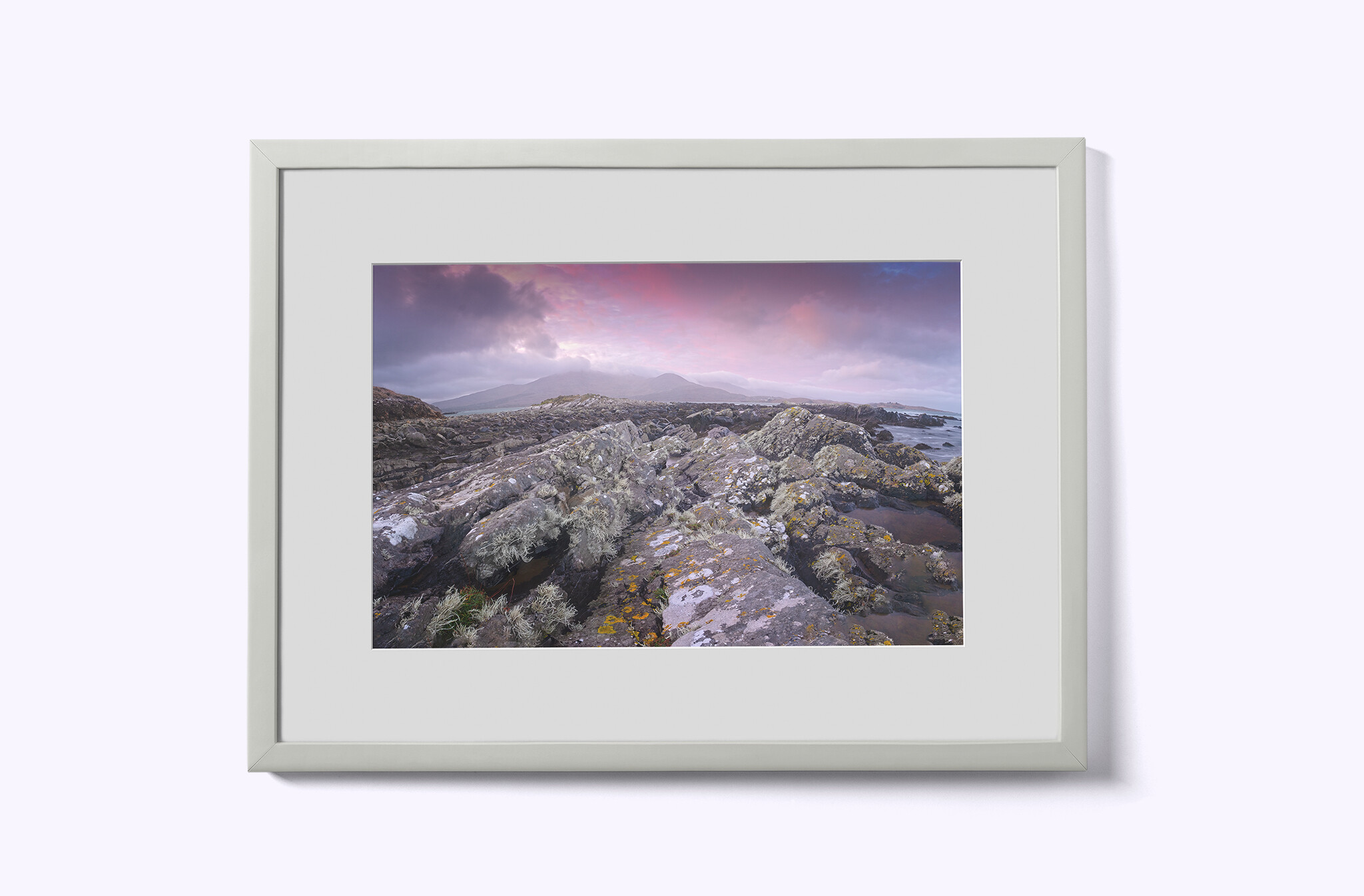 Framed photograph of a coastal landscape featuring rocky terrain, ocean waves, and distant mountains under a cloudy pink and purple sky. The frame is plain white, mounted against a light gray background.