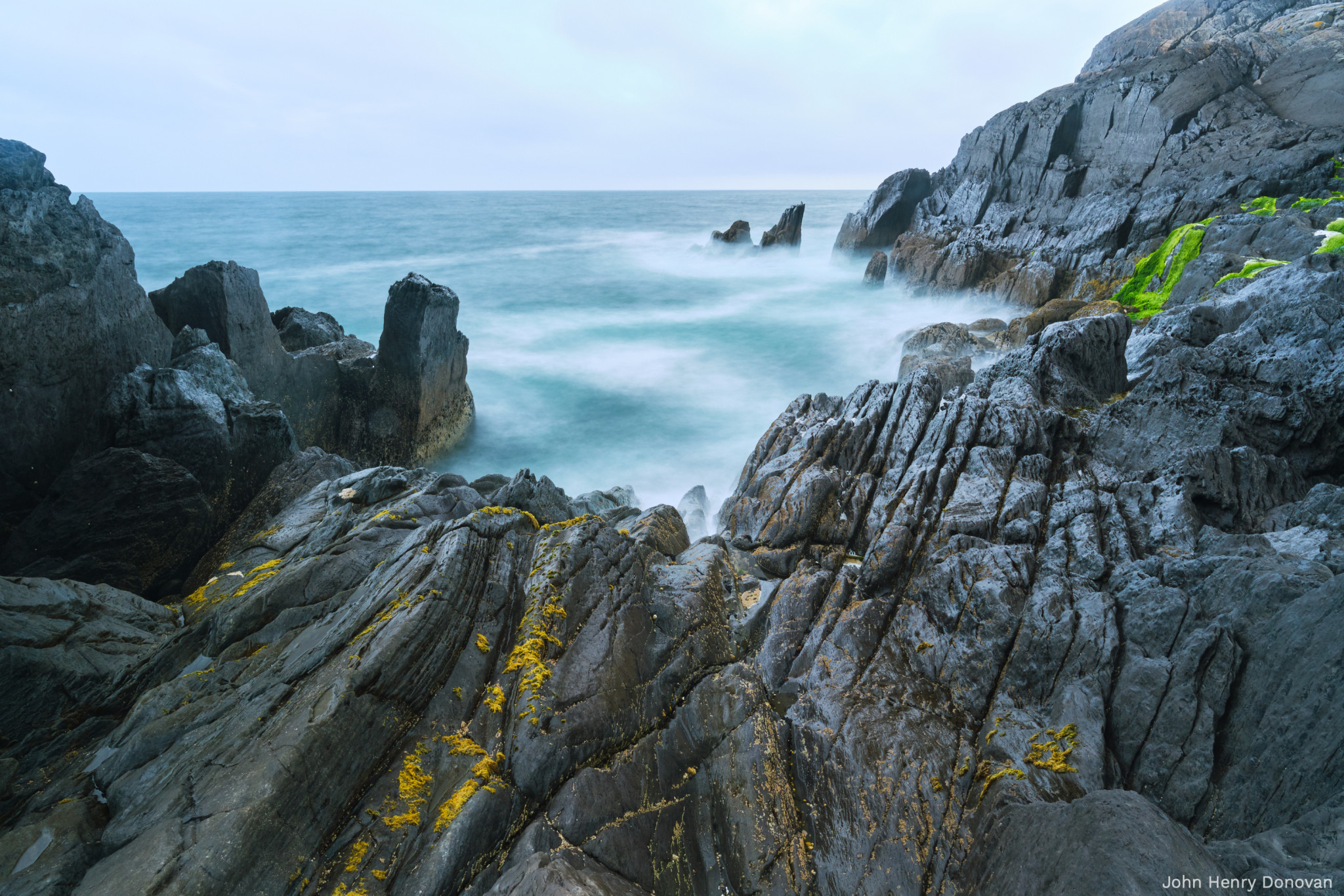 Rocky coastline with jagged rocks extending into the ocean. The sea appears calm with gentle waves, and some rocks in the foreground have yellow moss or seaweed on them. The sky is overcast, and there is a patch of green vegetation on the right.