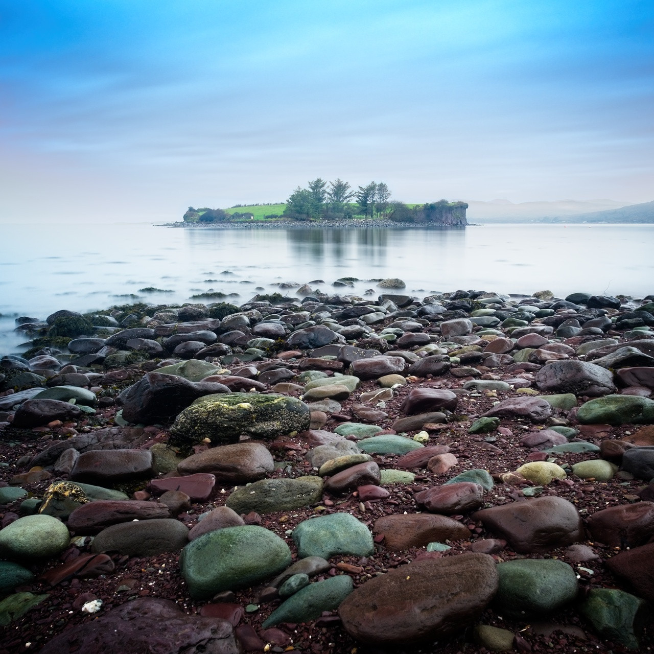 Rocky shoreline leading to a calm sea with a small island in the distance. The island is covered with green grass and a few trees. The sky is overcast, casting a bluish tone over the scene.