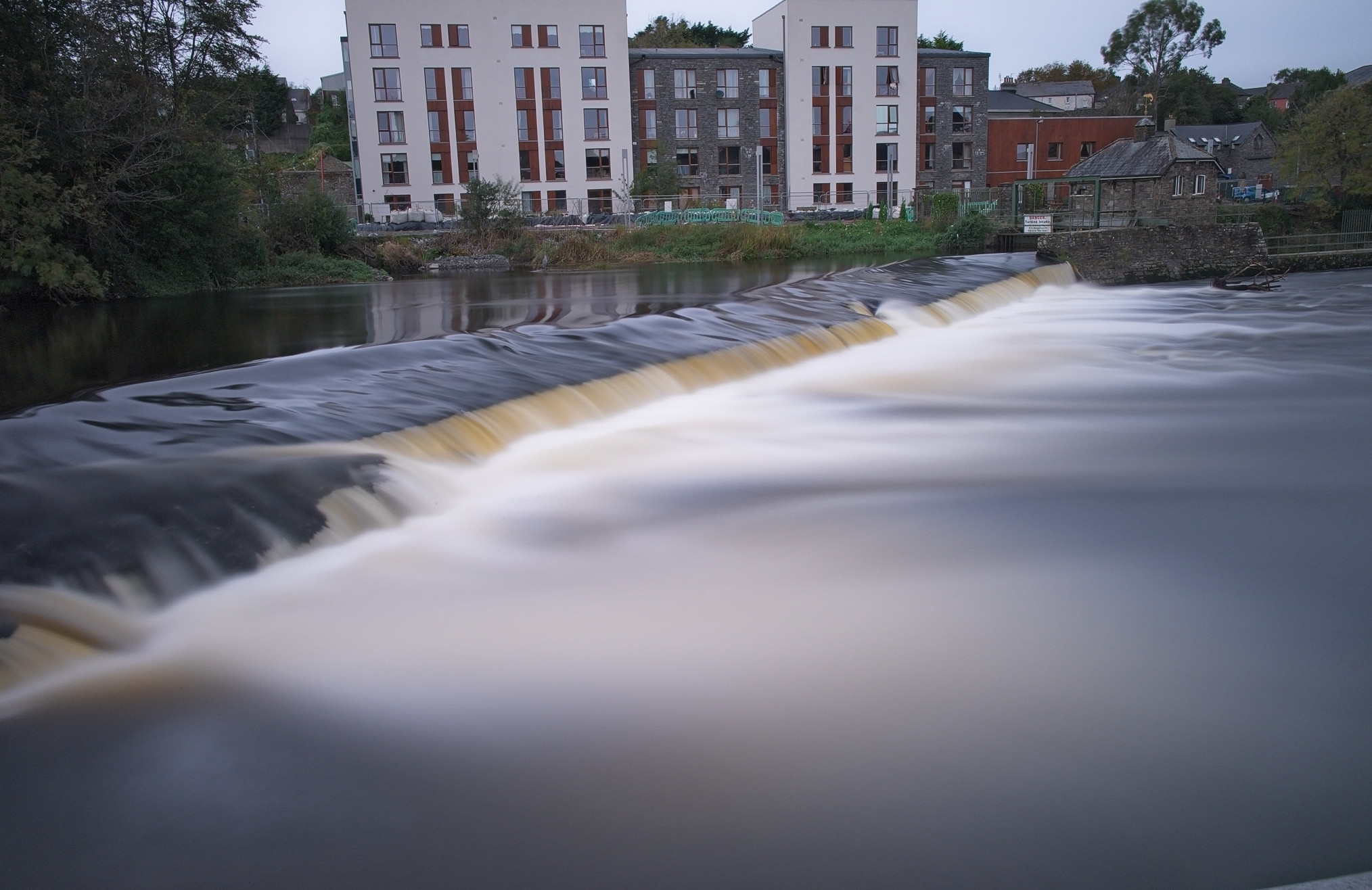 A wide, smooth waterfall flows over a weir on a river, with a modern, multi-story apartment building complex in the background. The adjacent area features greenery and a few older stone houses, suggesting a mix of urban and natural environments.