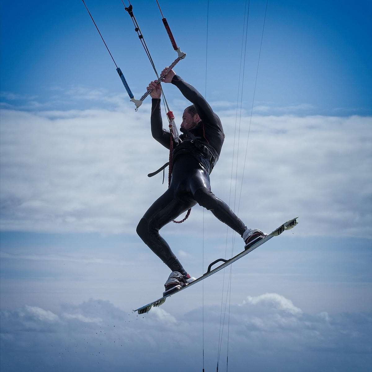 A person wearing a black wetsuit is kiteboarding against a backdrop of a cloudy sky. They are airborne, holding onto the kite control bar with both hands, and their legs are bent slightly at the knees. The person appears to be performing a jump or trick.
