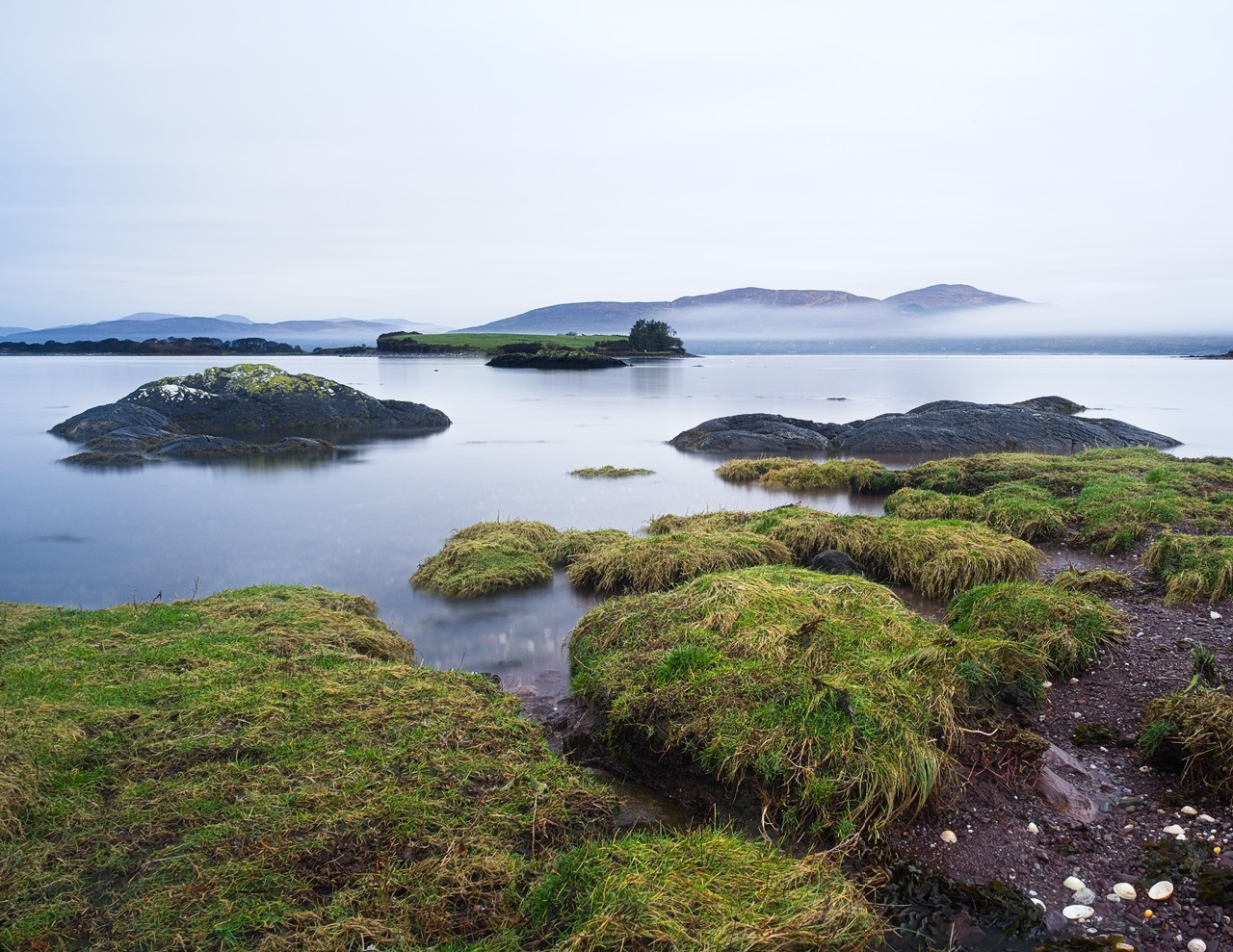 A calm body of water with small grassy islands and rocky outcrops. The scene is tranquil under a grey, overcast sky. In the distance, there are mist-covered mountains, adding a sense of depth and serenity to the landscape.