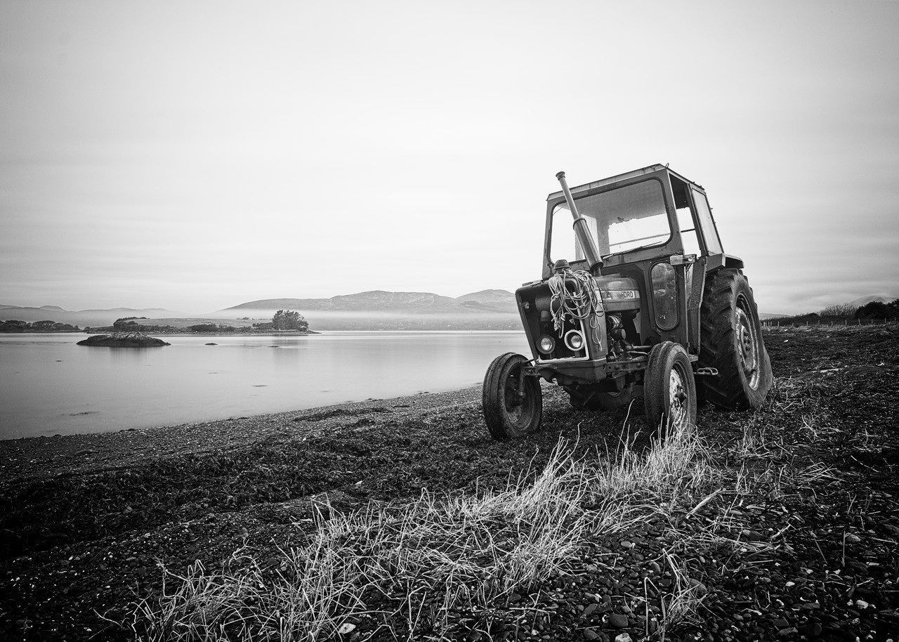 Black and white photo of a tractor parked on a grassy shore with a calm body of water and mountainous terrain in the background. The sky is overcast, and the tractor appears weathered. Small islands are visible in the distance.