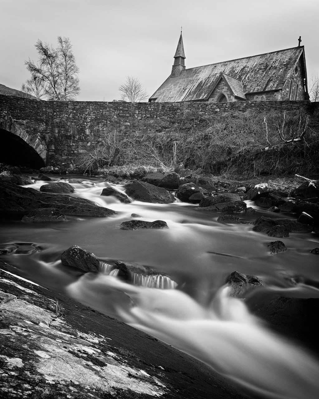 Black and white photograph of a serene landscape featuring a flowing stream with rocks in the foreground, a stone bridge above the stream, and a rustic church with a steeple in the background.