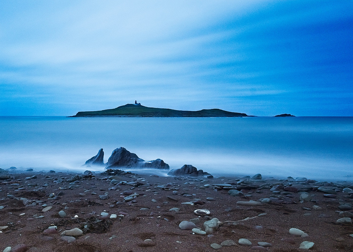 A calm seascape at dusk with a rocky shoreline in the foreground. In the distance, there is a small, flat island with a lone structure on it. The sky is mostly clear, with a light blue hue hinting at twilight.