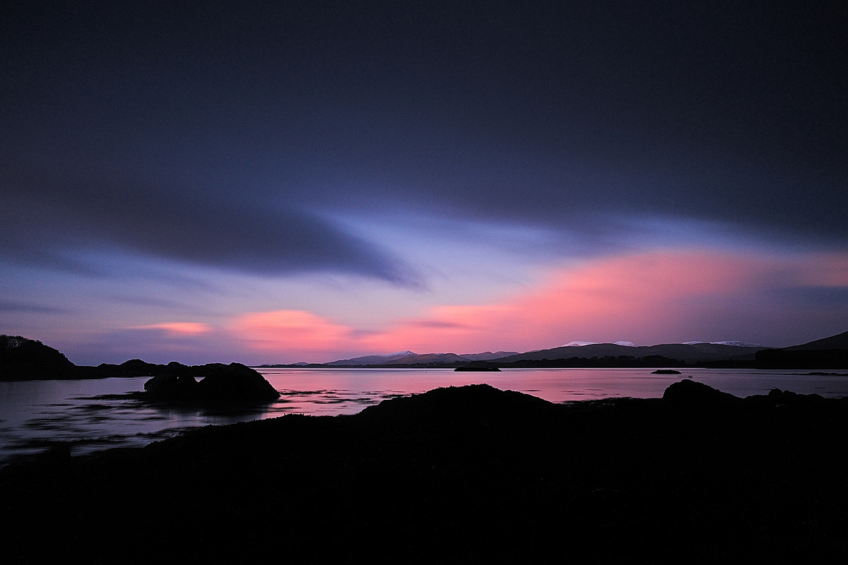 A tranquil coastal scene at dusk with a dark foreground contrasting a colorful sky. The sky transitions from deep blue to pink and purple hues, reflected in the calm waters. Rock formations and distant mountains are silhouetted against the horizon.