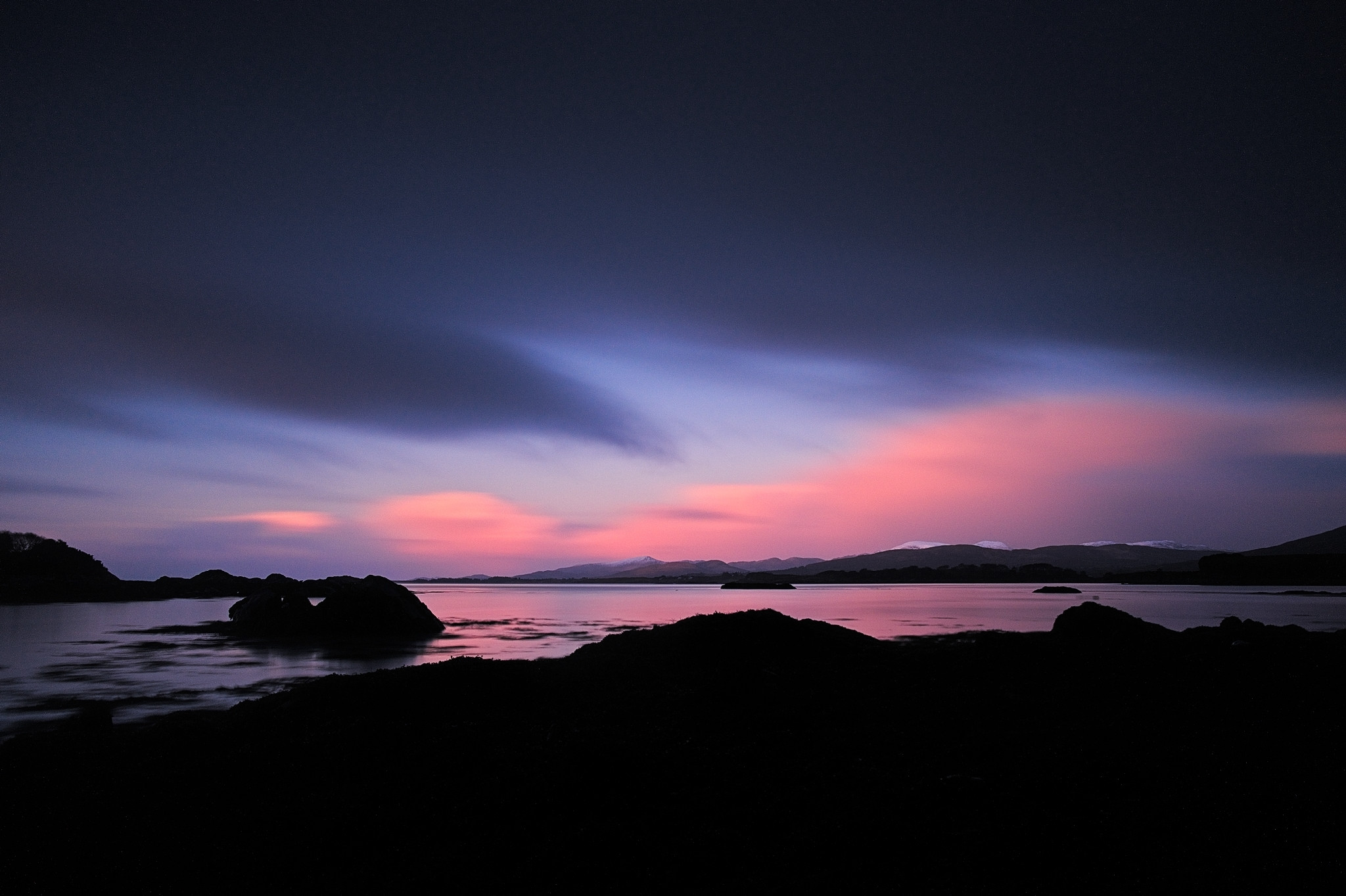 A tranquil coastal scene at dusk with a dark foreground contrasting a colorful sky. The sky transitions from deep blue to pink and purple hues, reflected in the calm waters. Rock formations and distant mountains are silhouetted against the horizon.