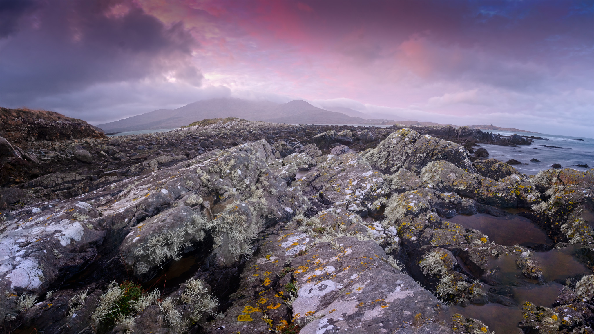 A rocky coastal landscape with rugged terrain covered in patches of lichen and moss. The ocean borders the rocks on the right, while a mountainous backdrop is visible under a purple and pink cloudy sky. The scene is tranquil and natural.