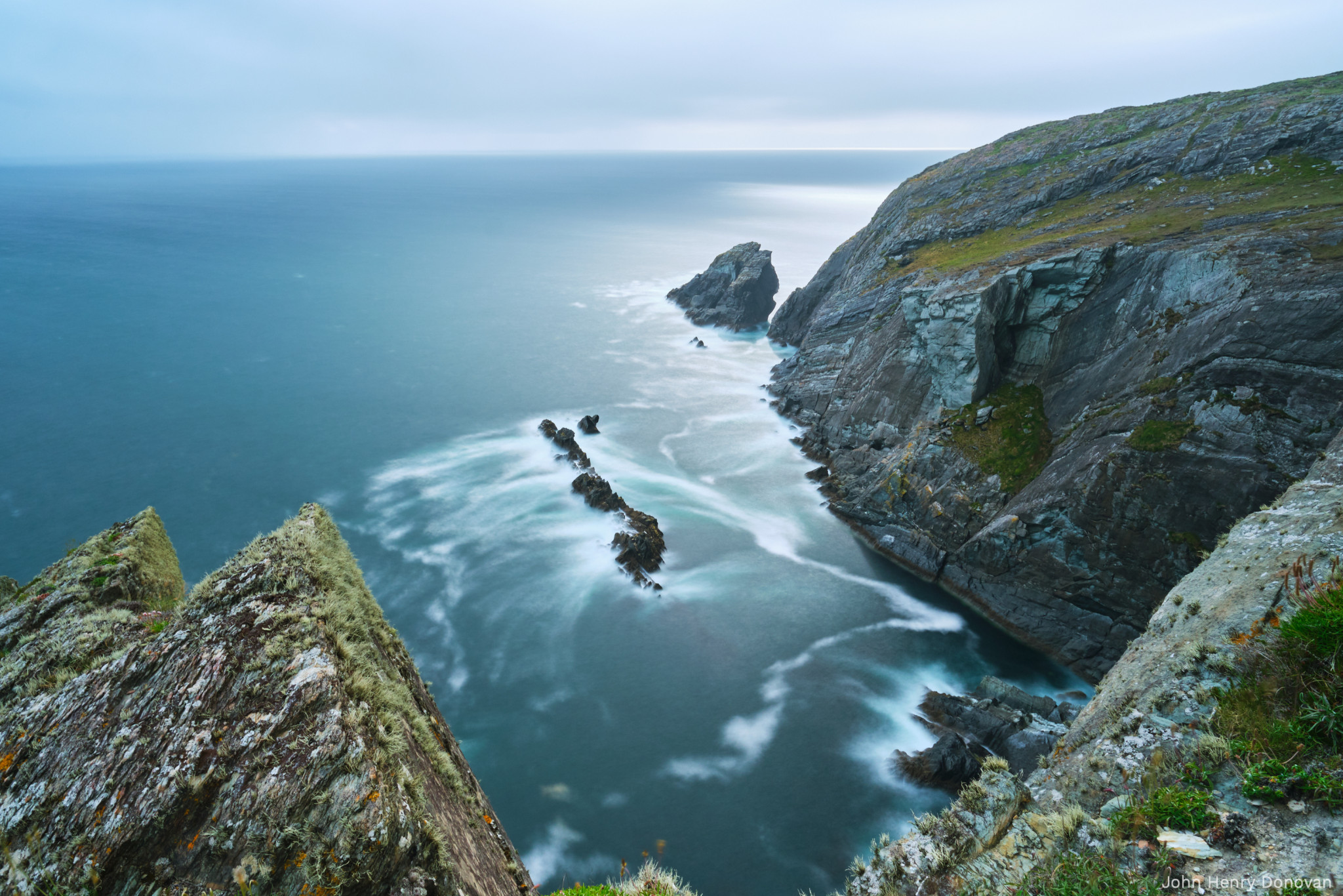 A coastal landscape shows rugged cliffs with greenery at the top, sloping into the ocean. The water appears smooth and misty, indicating long-exposure photography. Rock formations jut out into the sea, and a cloudy sky covers the horizon.