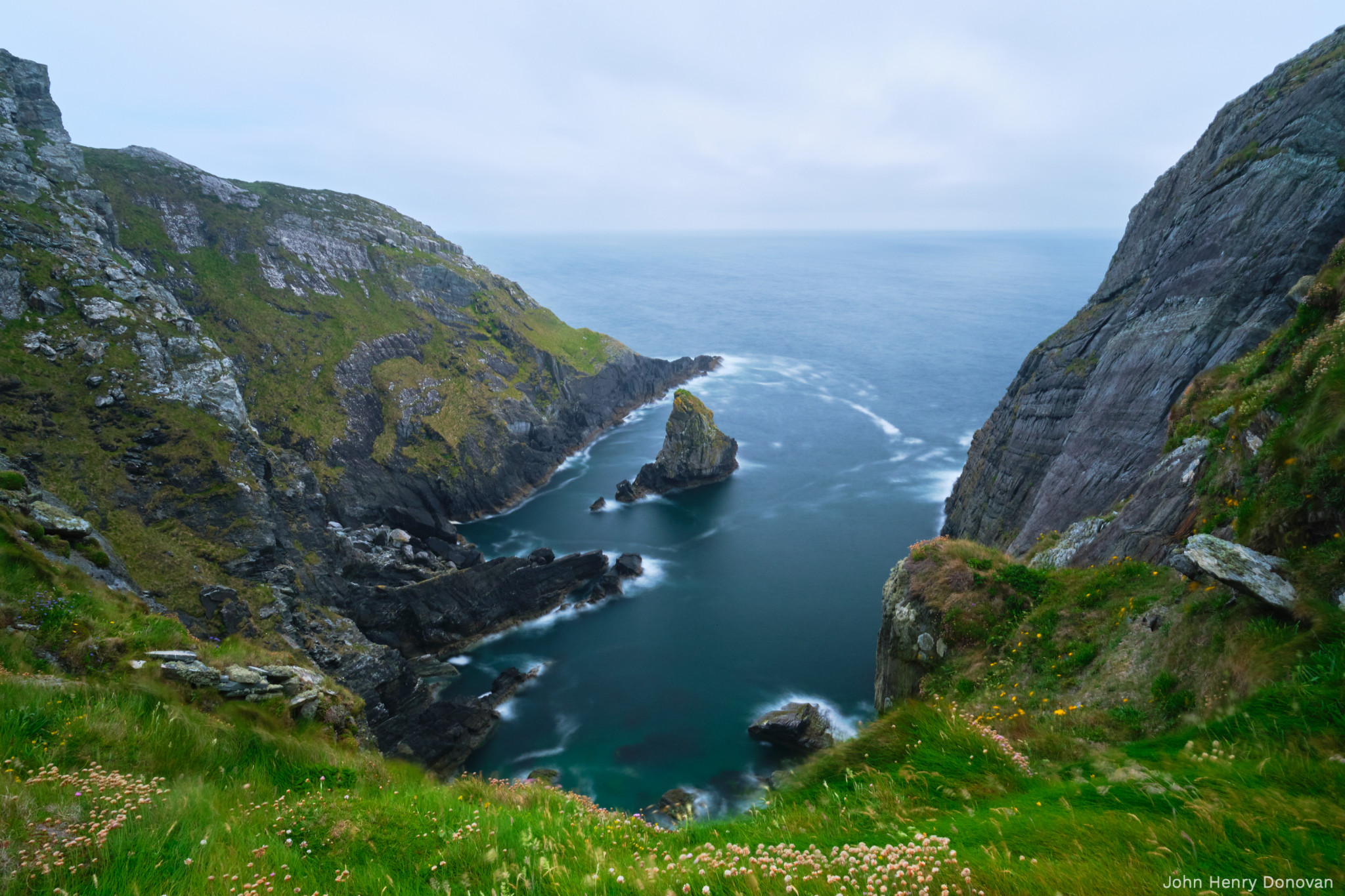 A coastal cliff scene with jagged rocks and a large boulder in the center of a narrow sea inlet. The grass-covered cliffs surround the inlet and extend into the ocean under an overcast sky. The water appears to be in motion with a blurred effect.