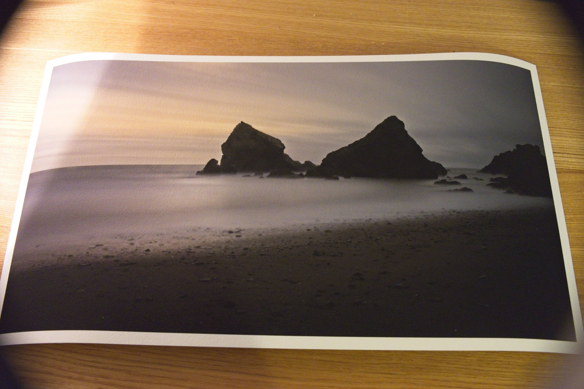 A printed photograph lies on a wooden surface. The image depicts a serene beach scene with dark, jagged rocks jutting out of calm ocean water under a cloudy sky. The photo features a long exposure effect, creating a smooth, misty appearance around the rocks.