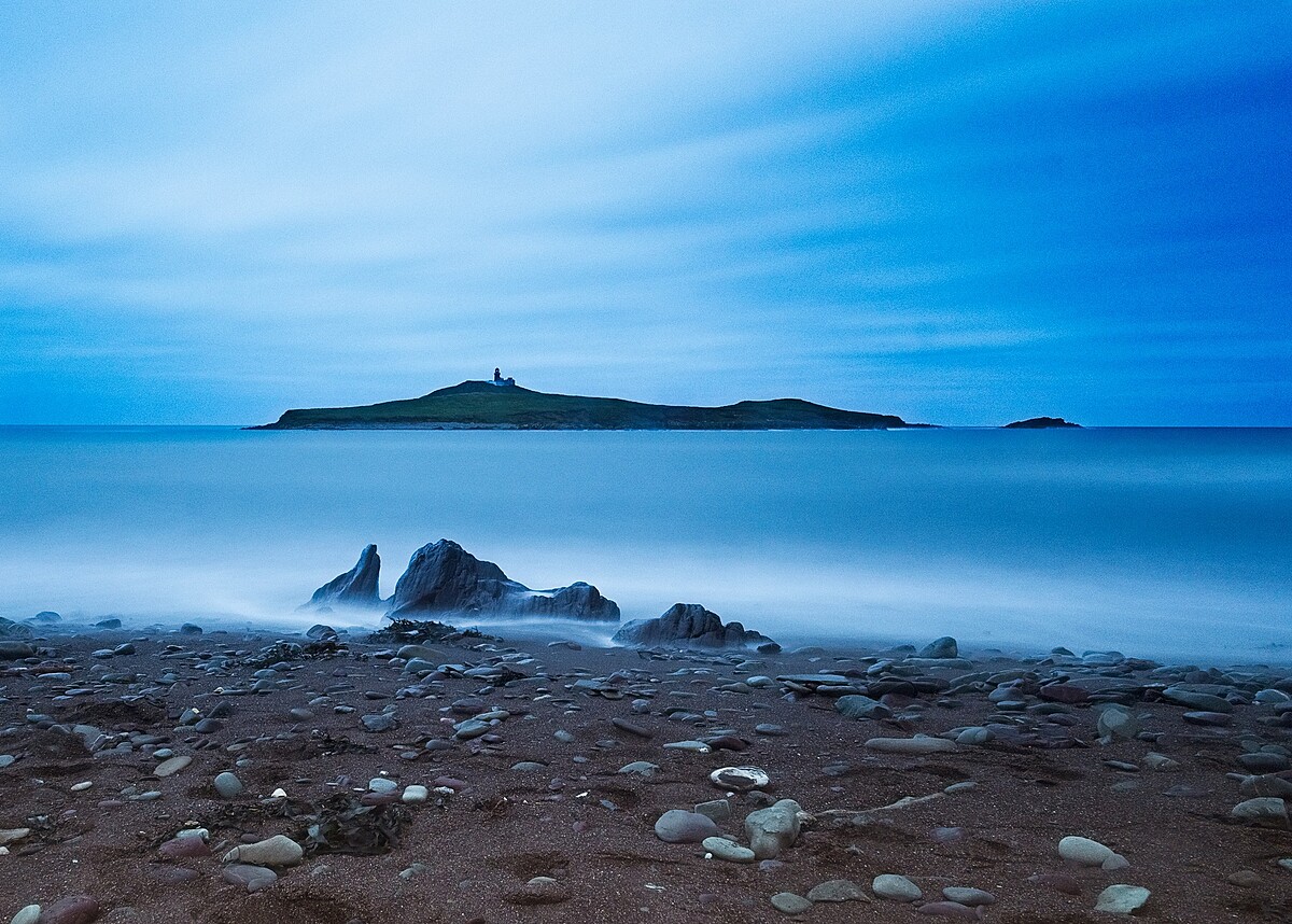 A calm seascape at dusk with a rocky shoreline in the foreground. In the distance, there is a small, flat island with a lone structure on it. The sky is mostly clear, with a light blue hue hinting at twilight.