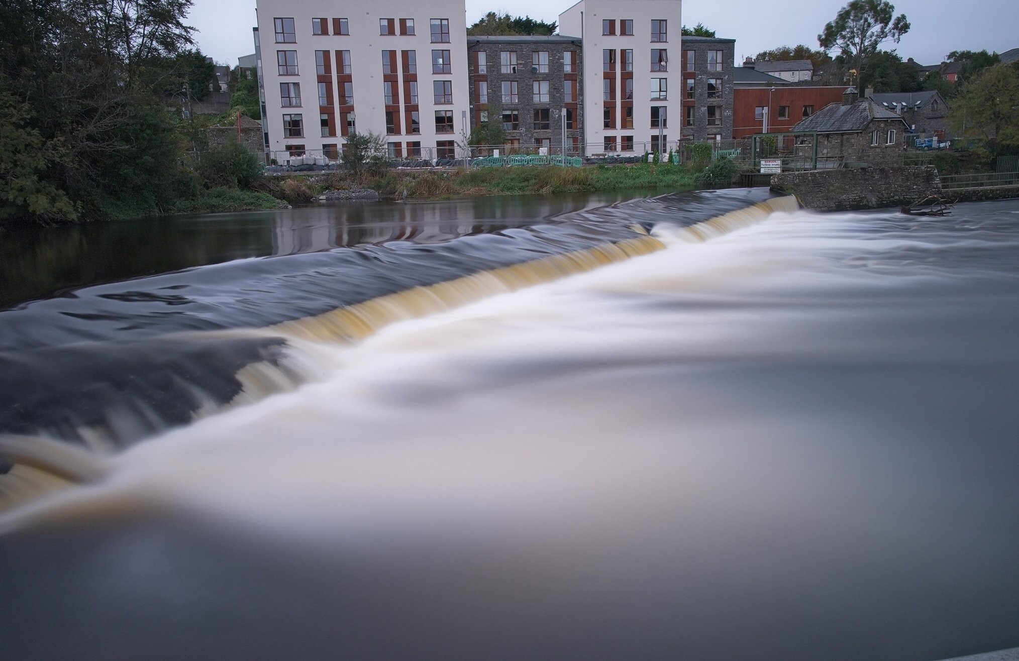 A wide, smooth waterfall flows over a weir on a river, with a modern, multi-story apartment building complex in the background. The adjacent area features greenery and a few older stone houses, suggesting a mix of urban and natural environments.